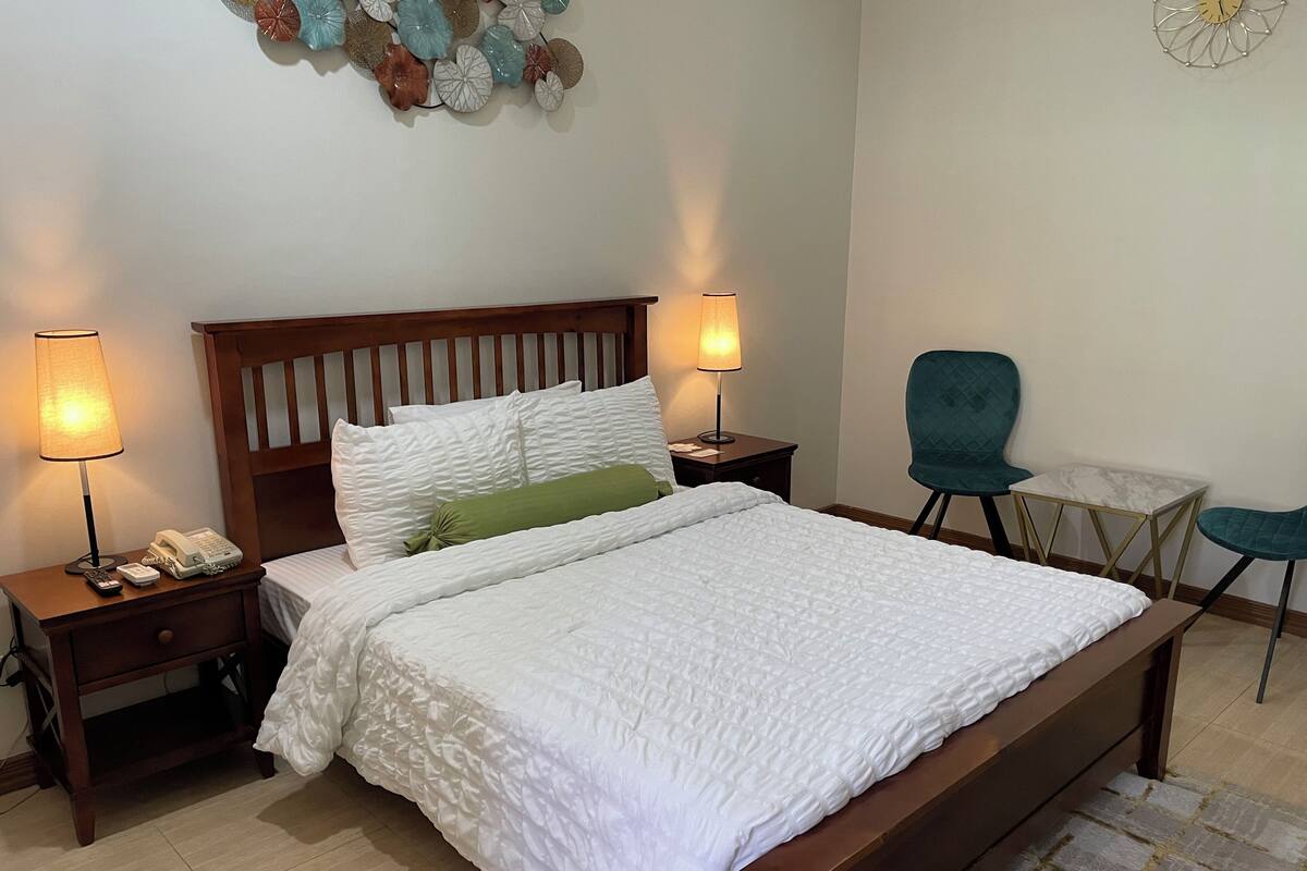 Un lit bien fait, recouvert d’une couette blanche, avec deux lampes de chevet, une table de chevet en bois, un oreiller vert et une petite table accompagnée d’une chaise.