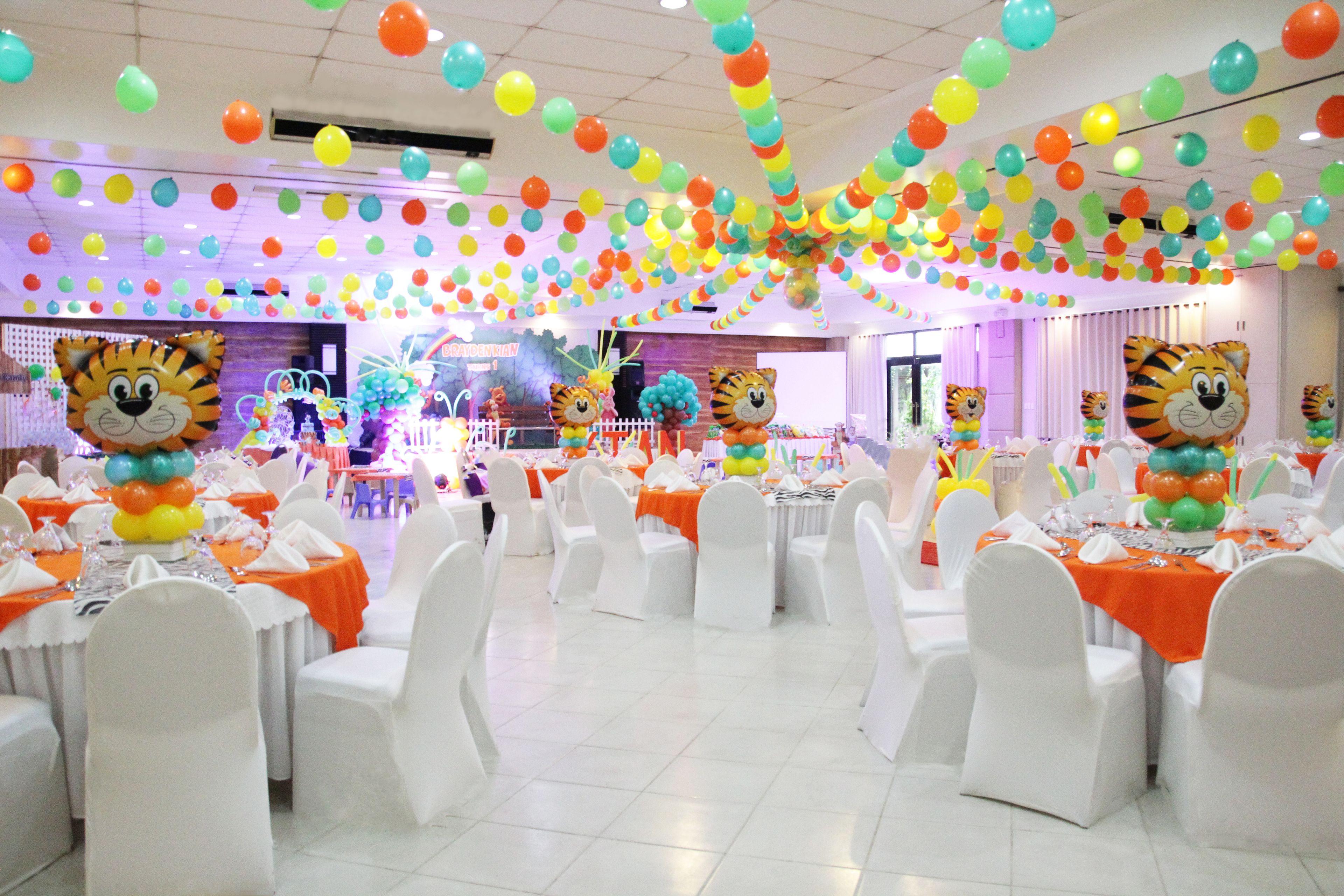 Un salón de banquetes con sillas y mesas blancas, decorado con globos y un gran adorno con forma de tigre.