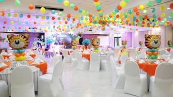 Un salón de banquetes con sillas y mesas blancas, decorado con globos y un gran adorno con forma de tigre.