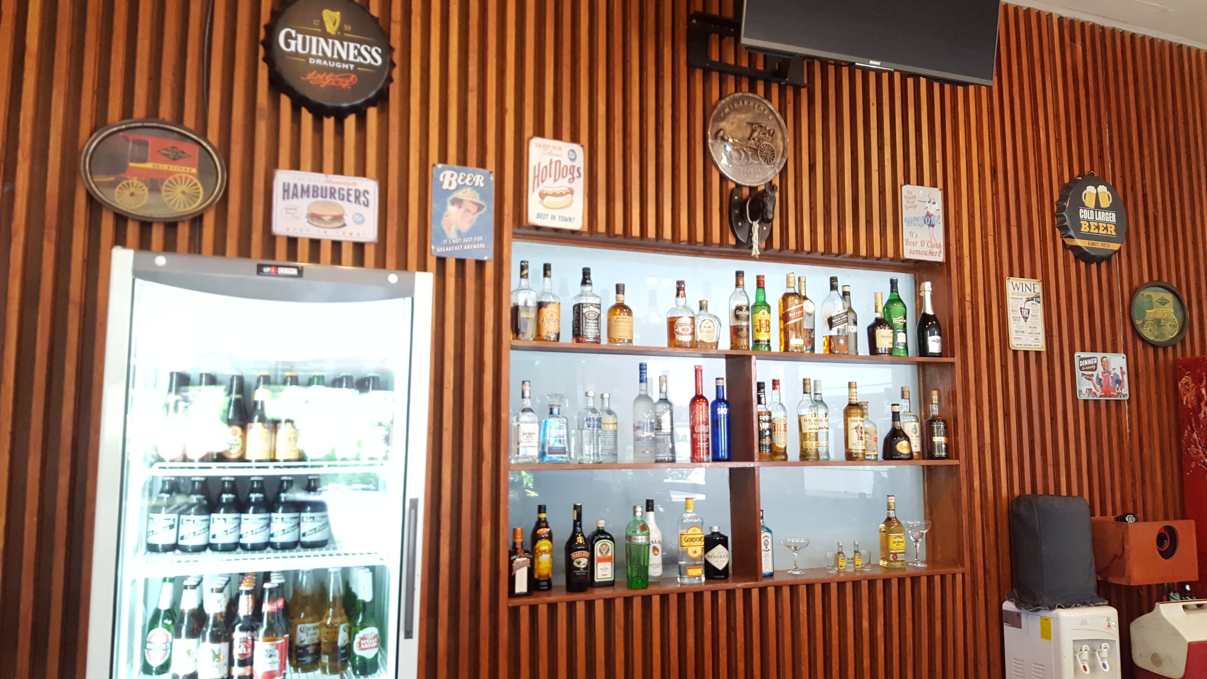 A bar area with a variety of liquor bottles, a refrigerator stocked with beverages, and several branded signs including Guinness and Hot Dogs.