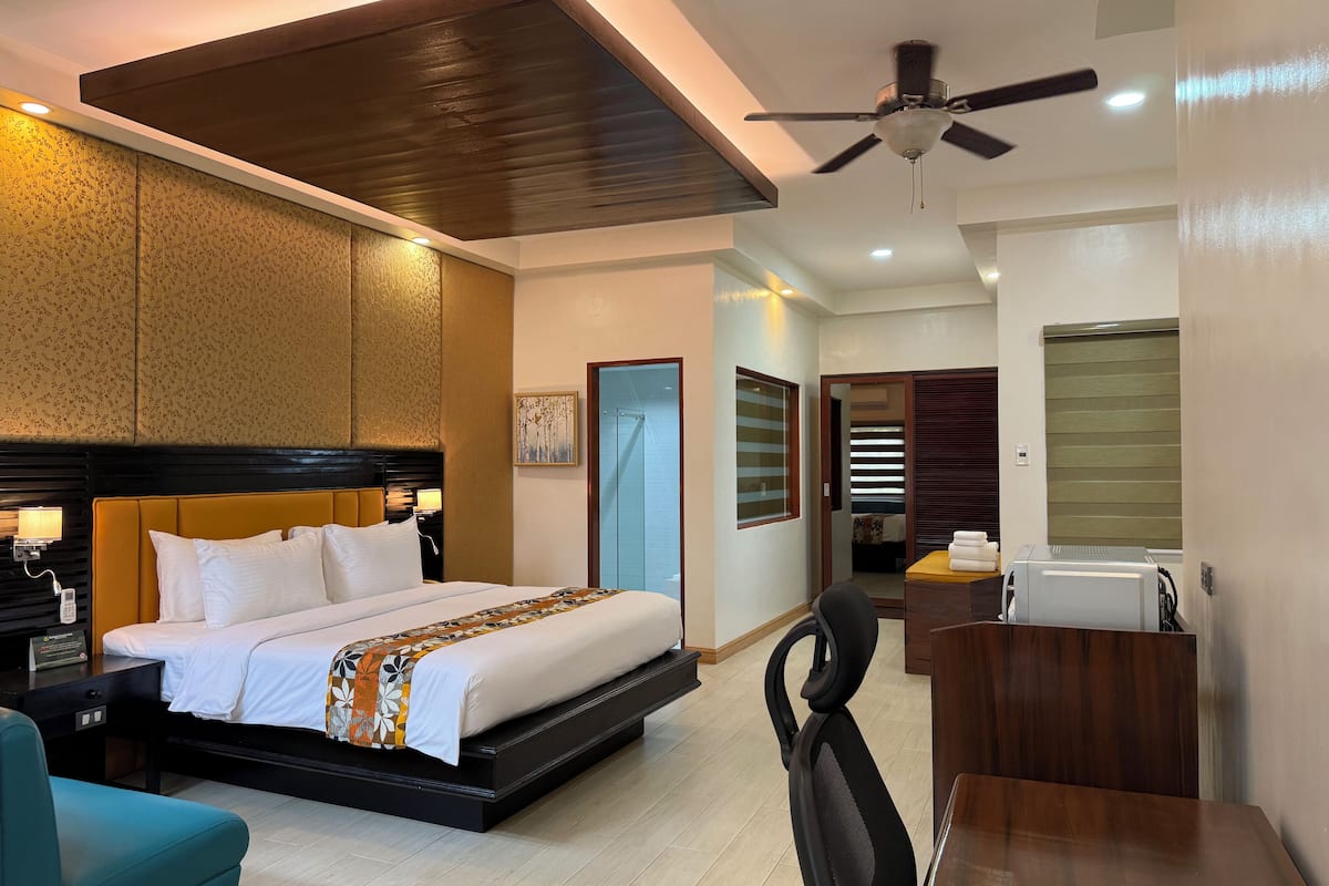 Une chambre d’hôtel avec un lit, un bureau, une chaise, un ventilateur de plafond et une salle de bain visible à travers une porte ouverte.