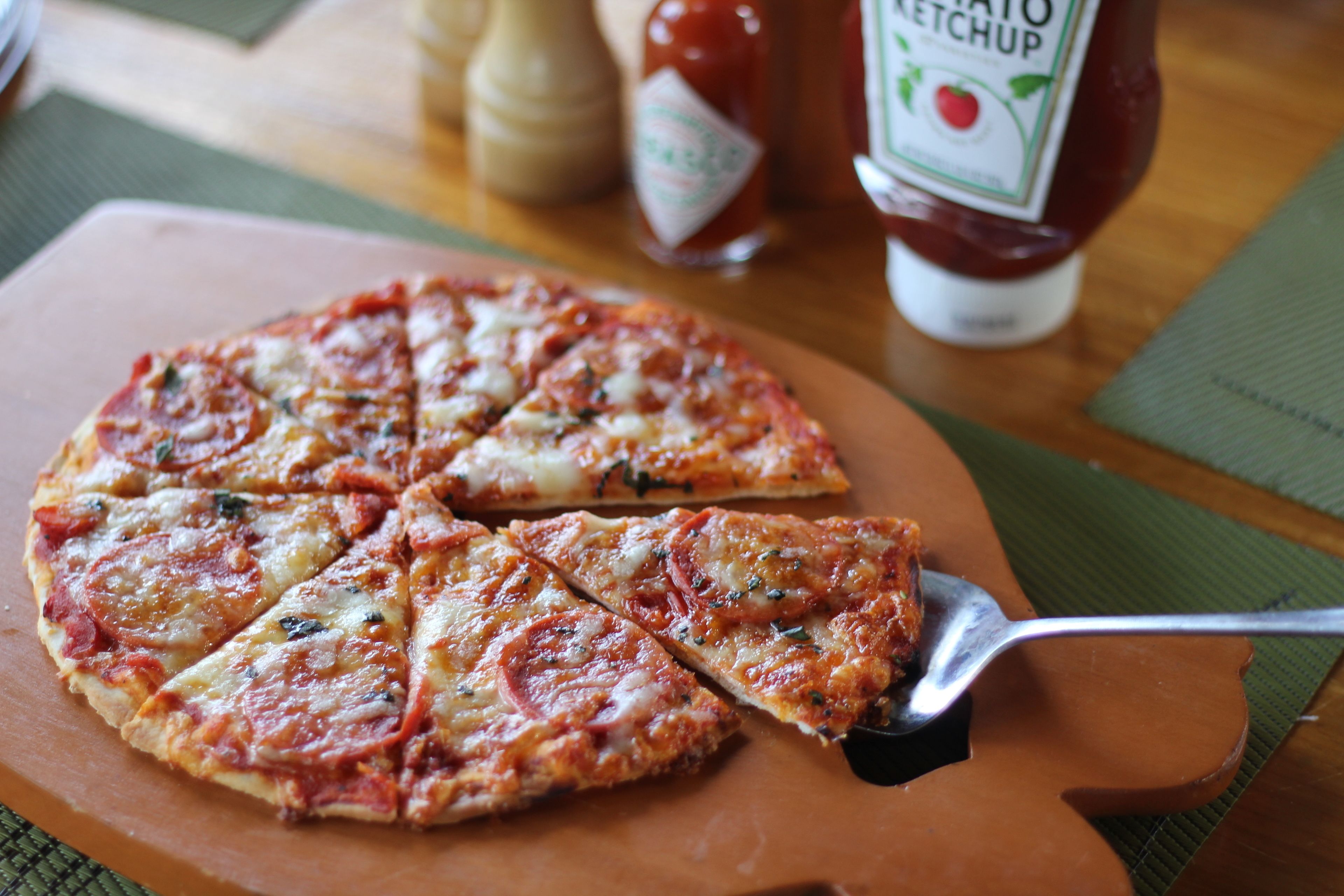 Eine Pizza auf einem Holzbrett, dazu eine Gabel. Im Hintergrund stehen Ketchup- und Senf-Flaschen.
