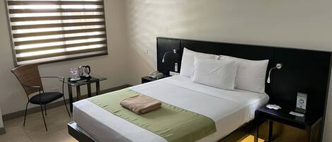 A hotel room with a large bed, a bedside table, a chair, a desk, and a window with blinds.