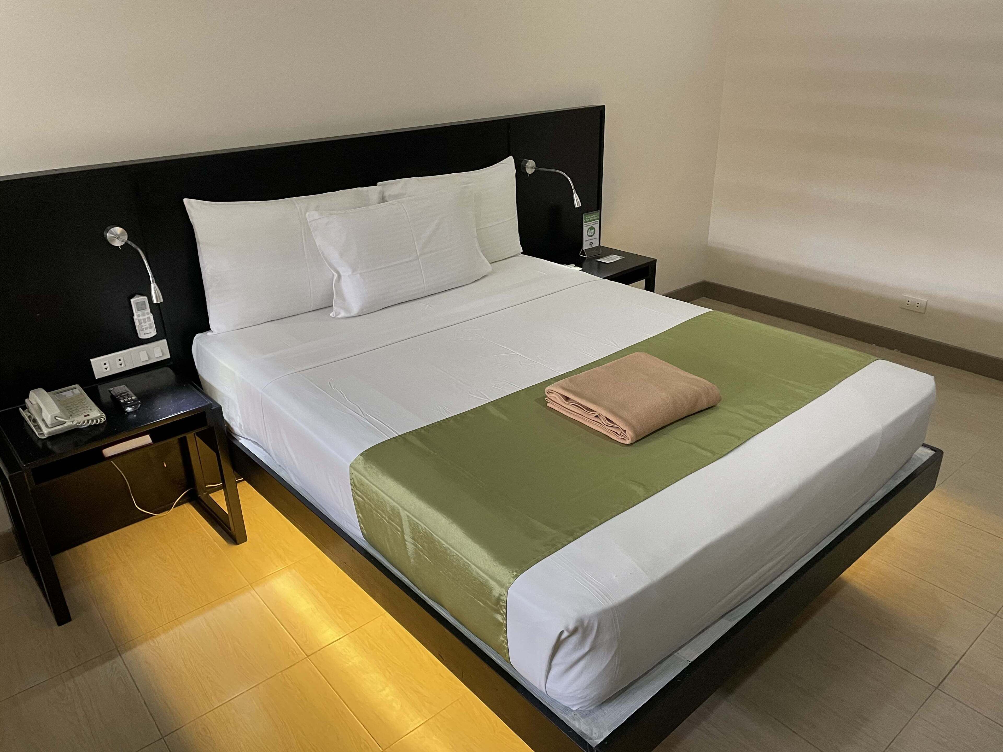 Cama bem arrumada com colcha verde e branca, toalha bege dobrada e almofadas brancas.  Há uma mesa de cabeceira com telefone e abajur. O quarto tem cabeceira preta e parede lisa.