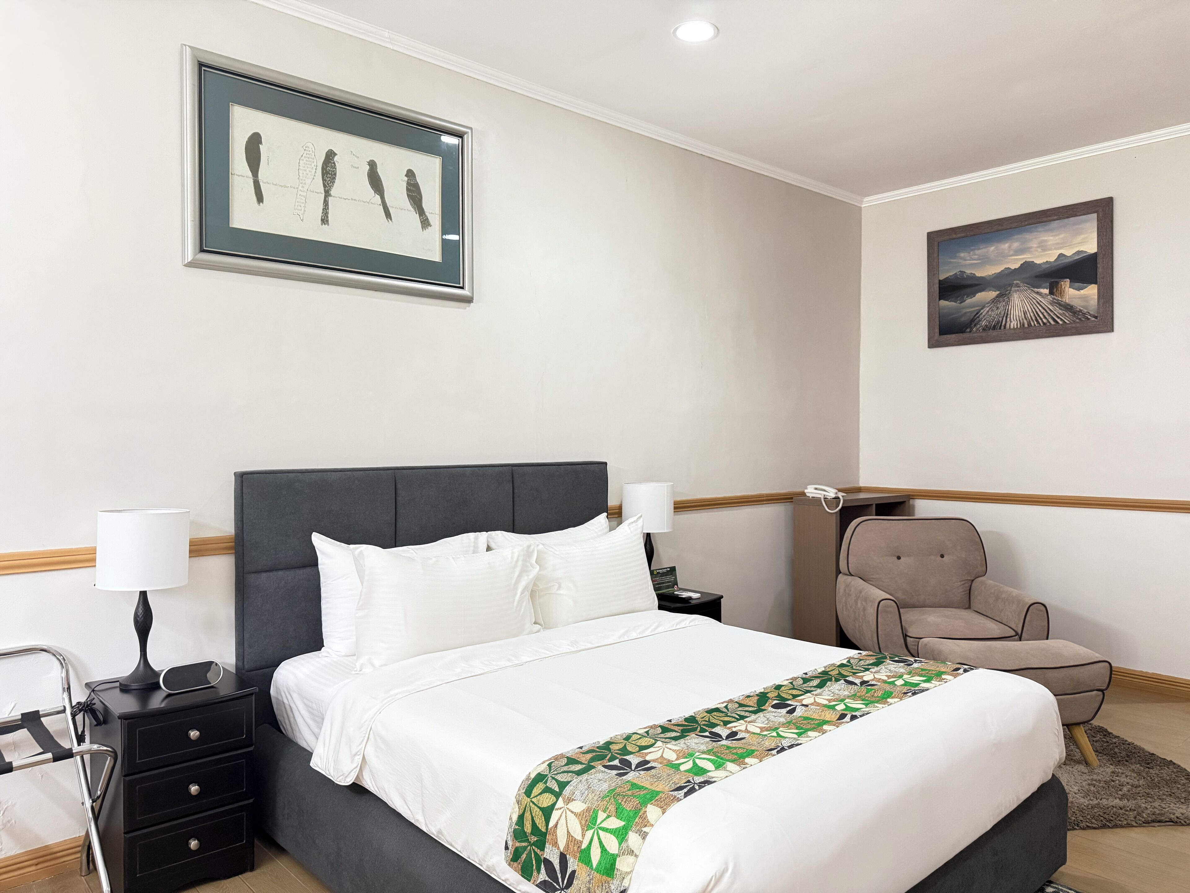 Habitación de hotel ordenada con una cama grande, mesitas de noche, una silla y dos cuadros en las paredes.
