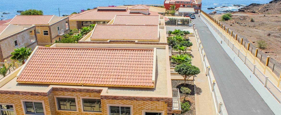 Zona residencial costera con edificios de tejas color terracota, una calle pavimentada y un litoral rocoso.