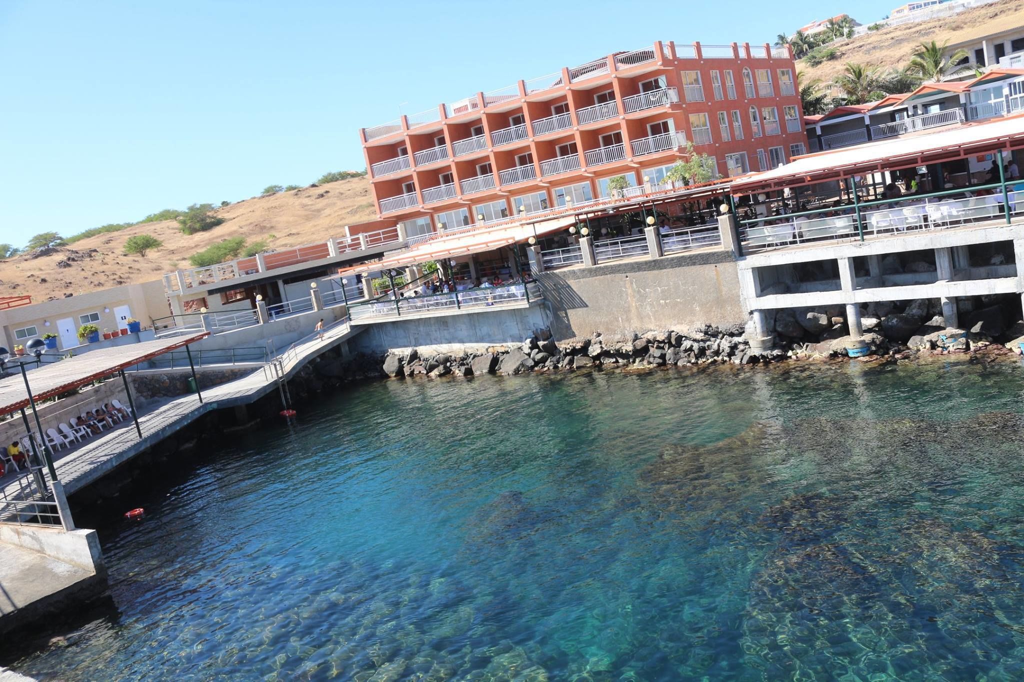 A waterfront building with balconies, a pier extending into the water, and a rocky shoreline.