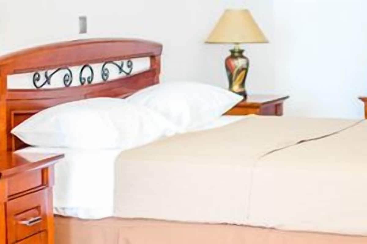 A neatly made bed with a wooden headboard, a bedside lamp, and a small bedside table with a book.