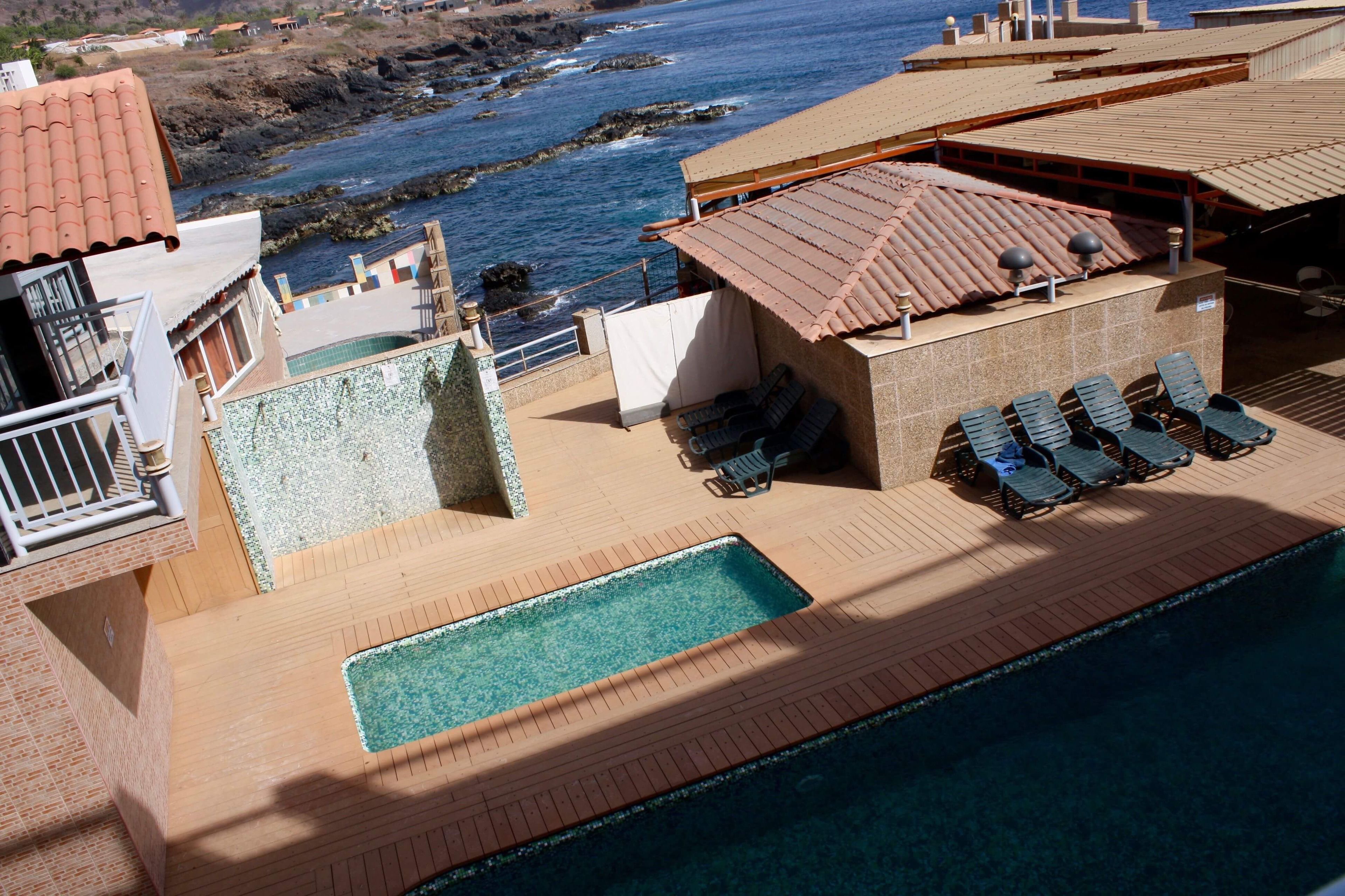 Un área de piscina en la azotea con vista al océano, sillones de descanso y un pequeño edificio con tejado de baldosas.