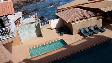 Un área de piscina en la azotea con vista al océano, sillones de descanso y un pequeño edificio con tejado de baldosas.