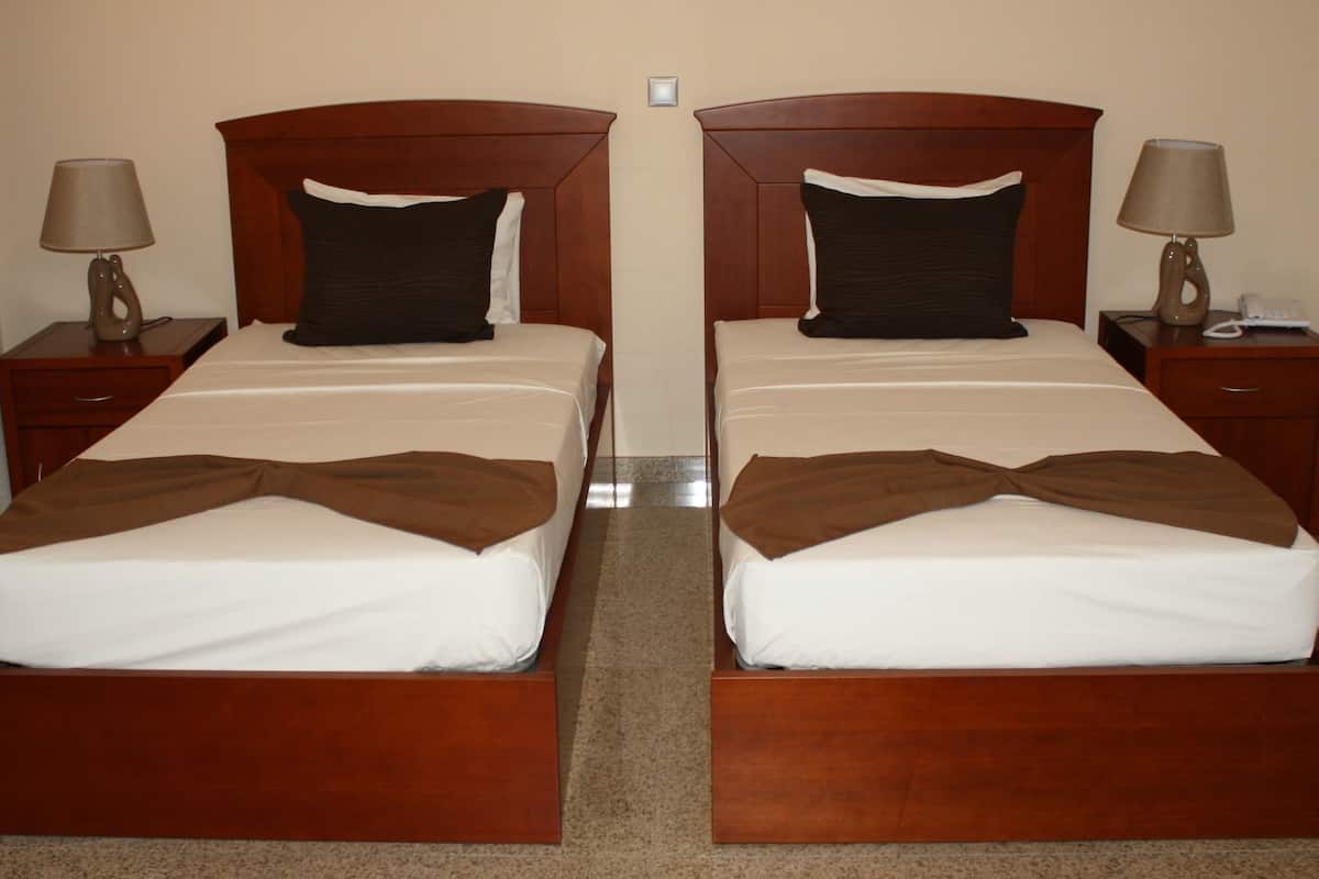 A hotel room with two identical beds, each with a wooden headboard, white linens, and a brown throw blanket. There are bedside tables with lamps and a telephone on each side.