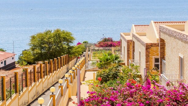 A coastal hotel with balconies overlooking the sea, a garden with pink flowers, and a view of the ocean. - Hotel Vulcão (Cidade Velha)