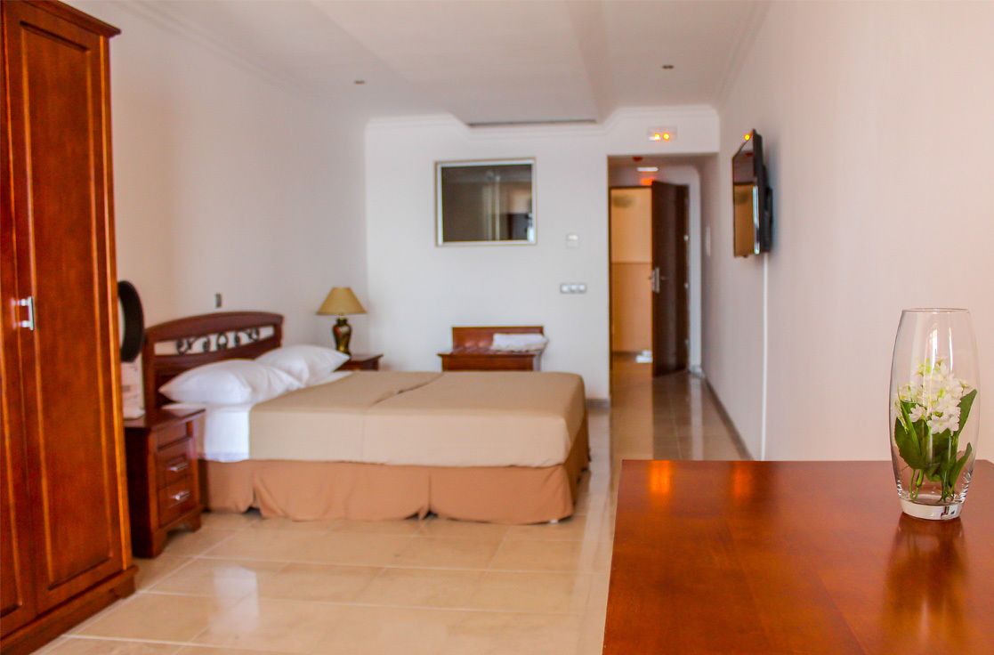 Una habitación de hotel con cama, armario de madera, mesita de noche, lámpara y un florero con flores.