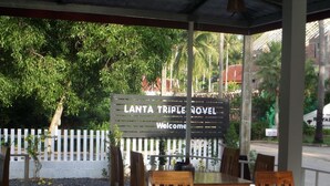 Un área de estar al aire libre techada con mesas y sillas de madera, un letrero que dice "LANTA TRIPLE NOVEL Bienvenidos" y una valla de piquetes blancos al fondo.