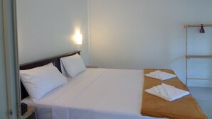 Una cama individual con sábanas blancas, cabecera de madera y una mesita de noche con lámpara. Hay una mesa de madera con toallas dobladas y un pequeño estante con un objeto morado.