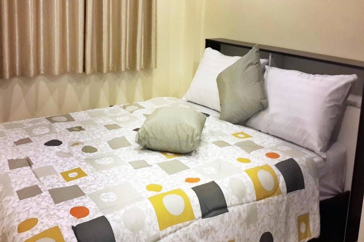 A neatly made bed with a patterned bedspread and pillows, a headboard, and a window with curtains.