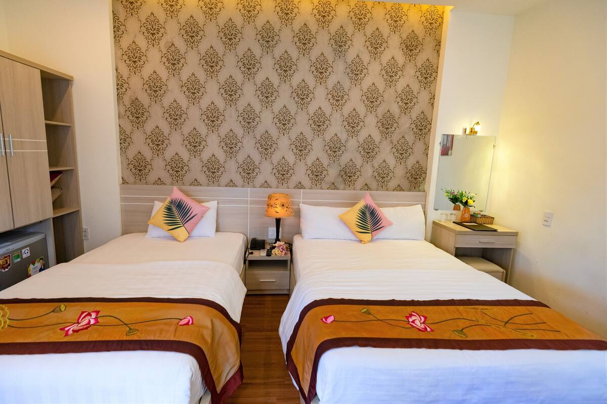 Habitación de hotel con dos camas, un armario, una mesita de noche con lámpara y una pequeña mesa con una planta.