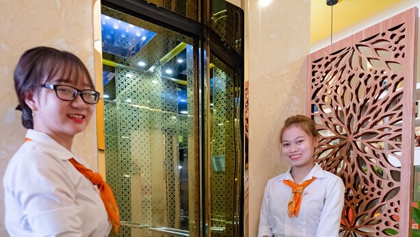Two individuals standing in a lobby with a glass door, a decorative wooden panel, and a tiled floor. - Flora Hotel Hue (Hue)