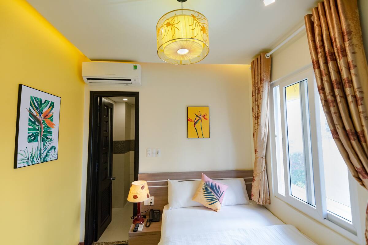 Habitación de hotel con cama, mesita de noche, lámpara, aire acondicionado y ventana con cortinas.
