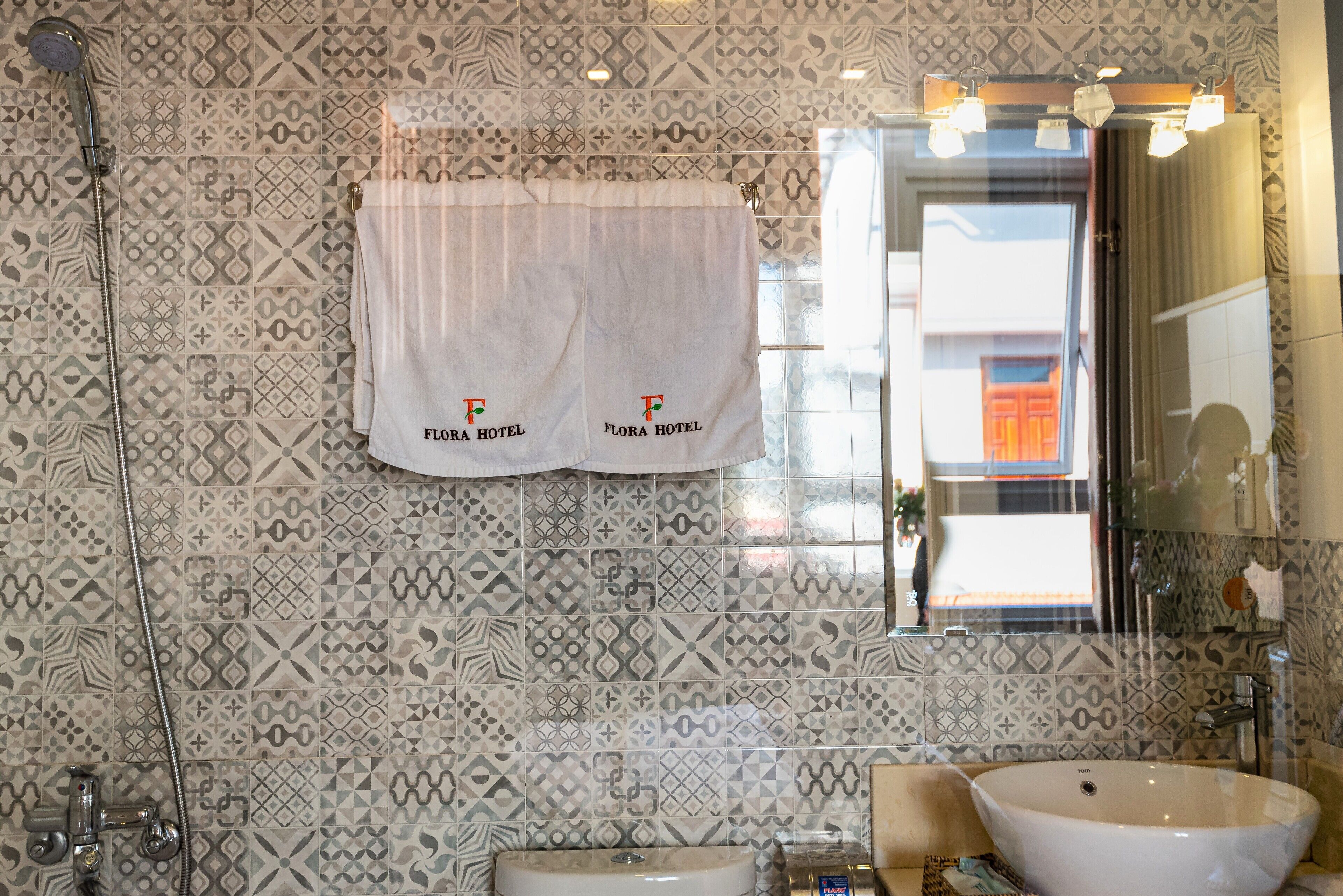 A bathroom with patterned tiles, a white towel with 'Flora Hotel' branding, a sink, and a shower.