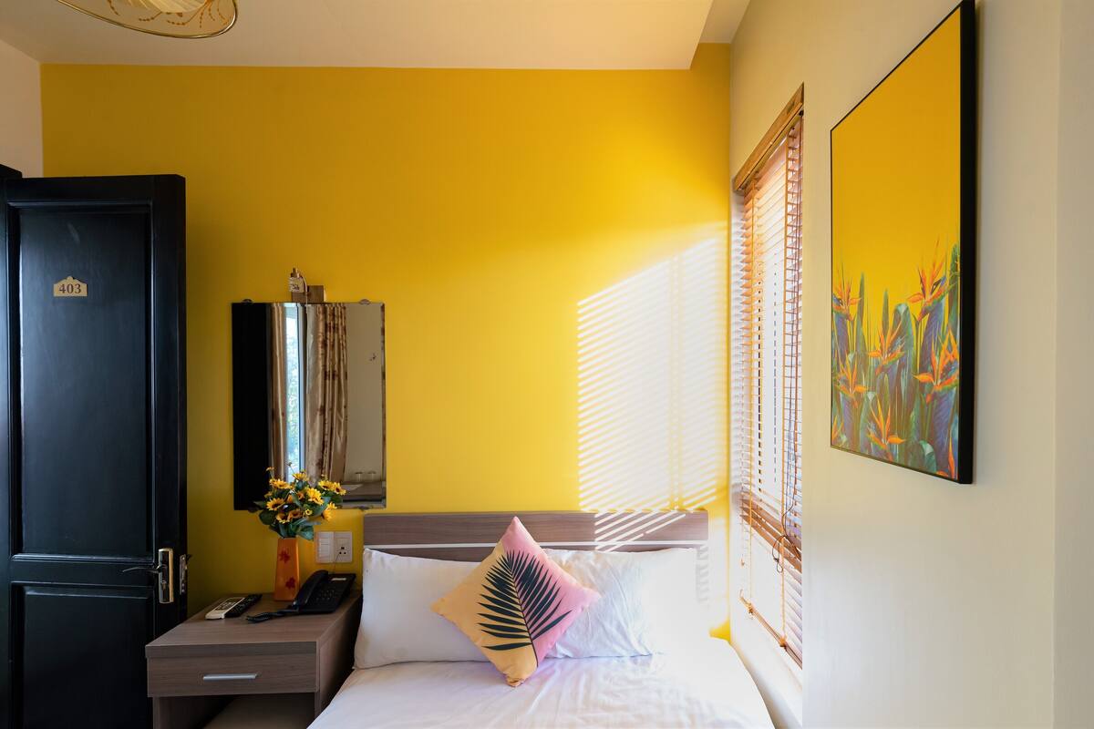 Habitación de hotel con cama, escritorio, teléfono, un florero con flores y un cuadro en la pared.