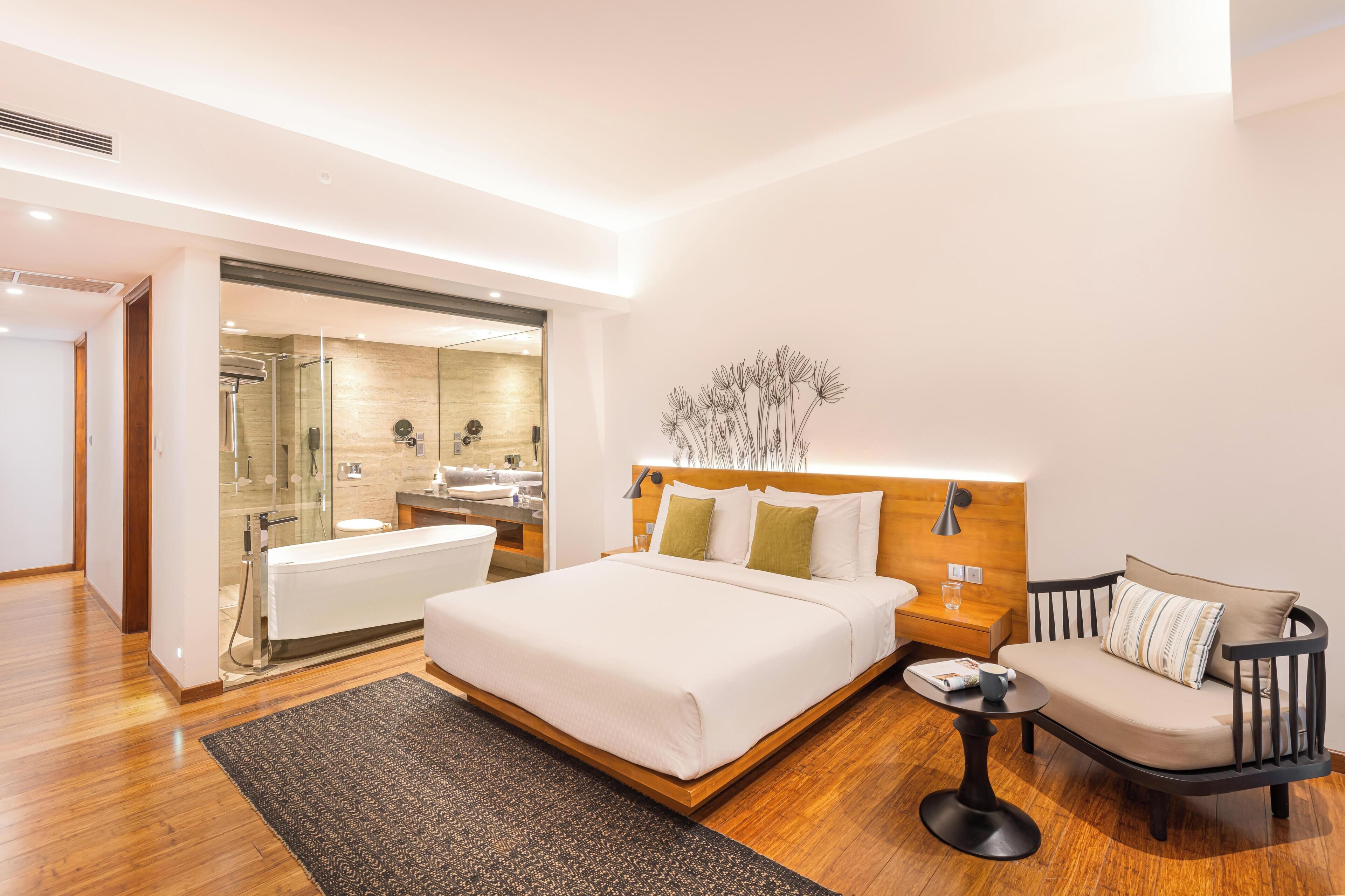 Ein modernes Hotelzimmer mit einem großen Bett, einem Nachttisch, einem Sessel, einem kleinen Tisch, einem Spiegel und einem Badezimmer mit Dusche und Waschbecken.