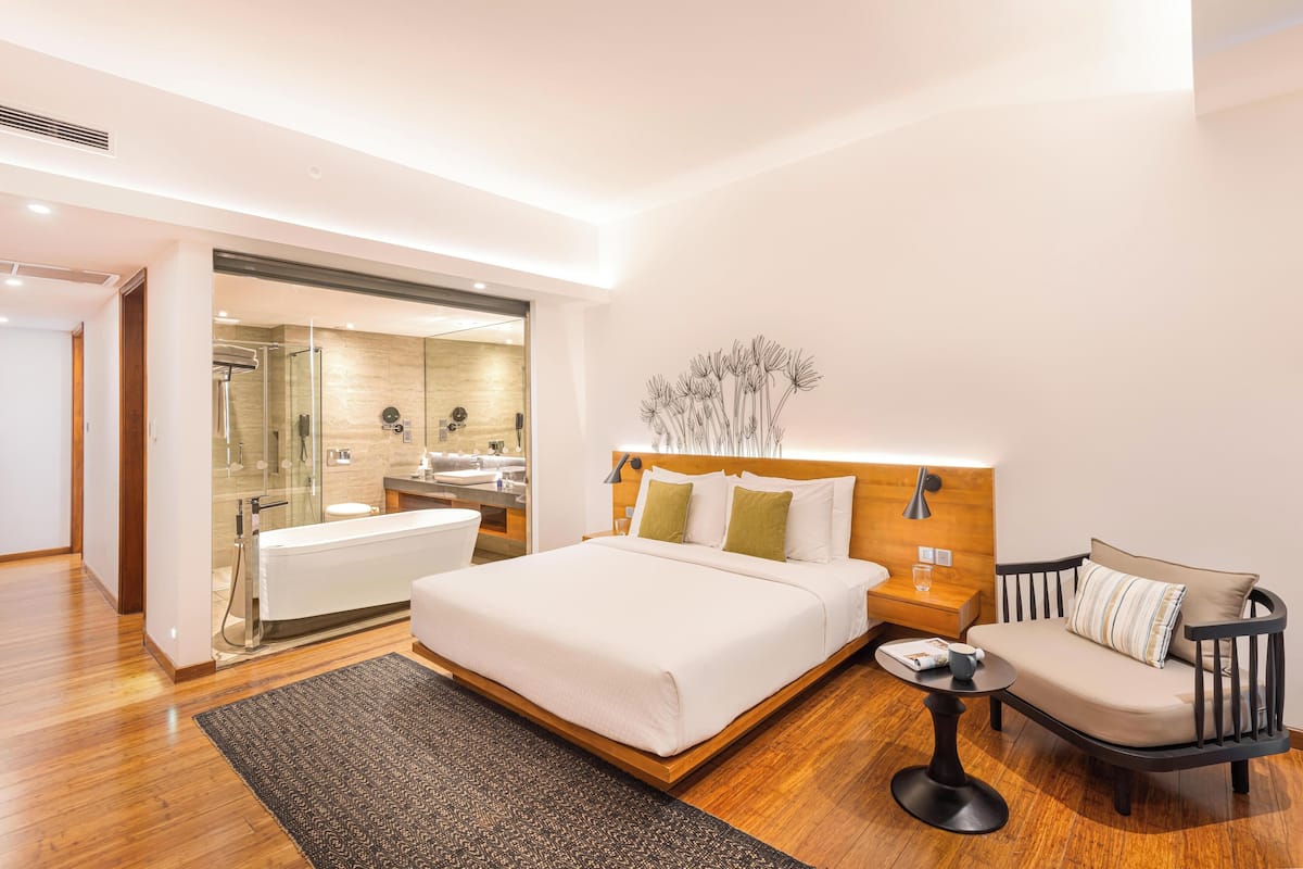 Une chambre d’hôtel moderne dotée d’un grand lit, d’une table de chevet, d’une chaise, d’une petite table et d’une salle de bain avec une douche à parois de verre.
