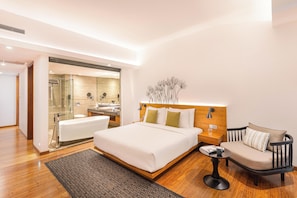 A modern hotel room with a large bed, a bedside table, a chair, a small table, and a bathroom with a glass shower enclosure.