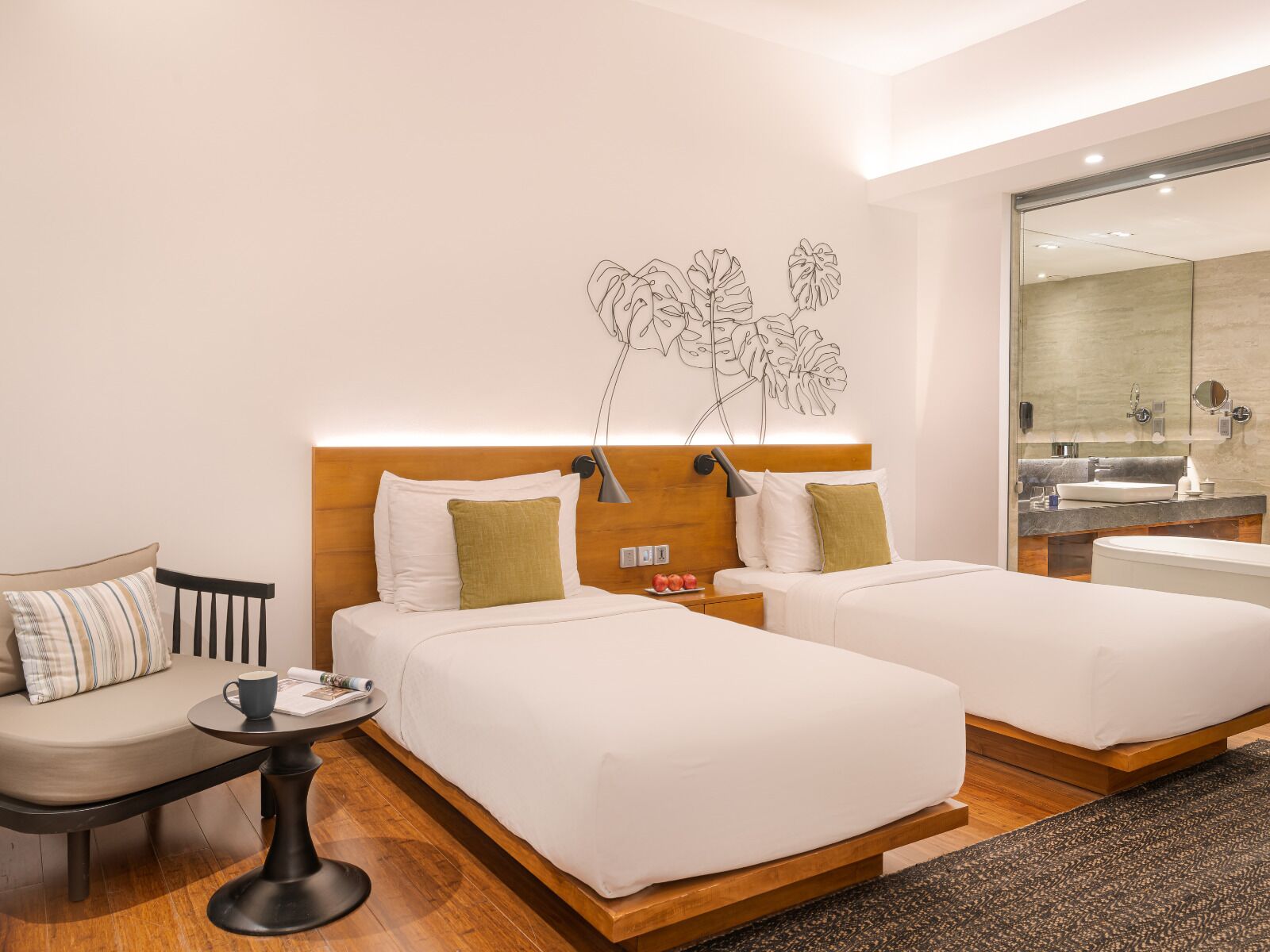 A hotel room with two beds, a small table with a cup, a chair, a wall art of leaves, and a bathroom visible through a glass door.