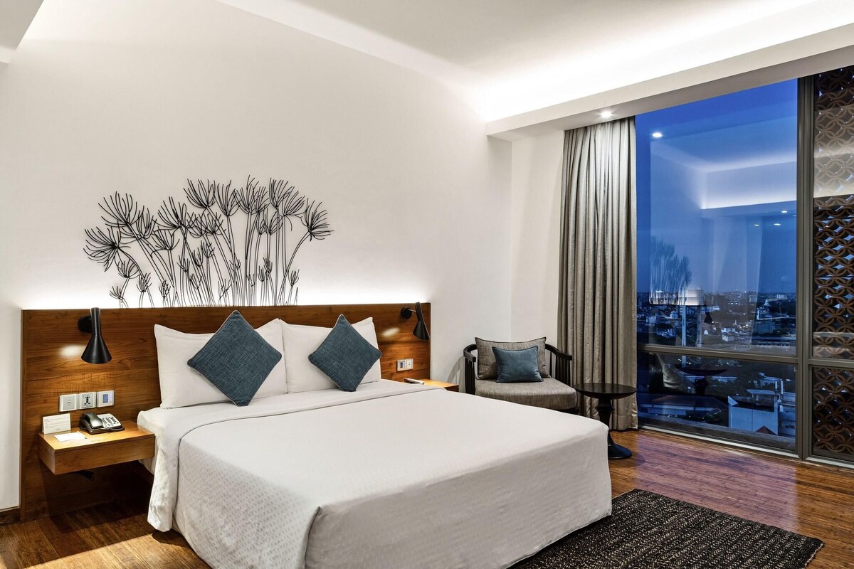 Quarto de hotel moderno com uma cama grande, uma mesa de cabeceira com telefone, uma luminária na parede e vista da cidade por grandes janelas.