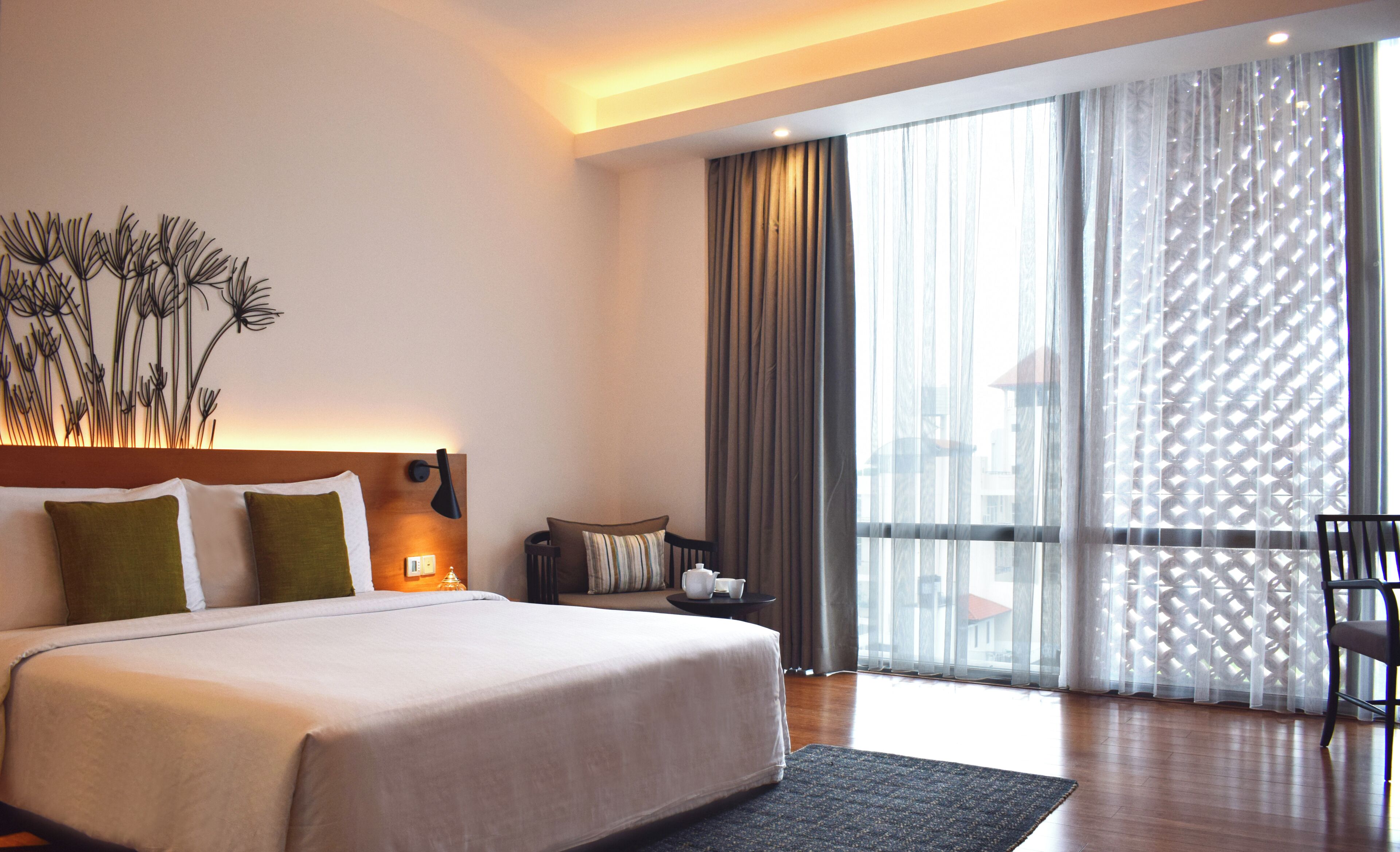 A modern hotel room with a large bed, a nightstand, a chair, and a view of the city through sheer curtains.