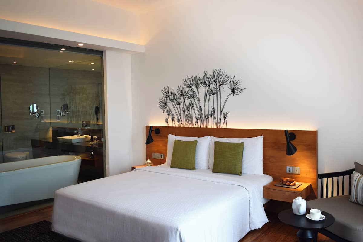 Une chambre d’hôtel moderne avec un grand lit, une table de chevet avec une lampe, une petite table ronde avec une théière et une tasse, et un canapé avec un coussin.