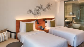 A hotel room with two beds, a wooden headboard, a wall-mounted light fixture, and a bathroom visible through a glass partition.