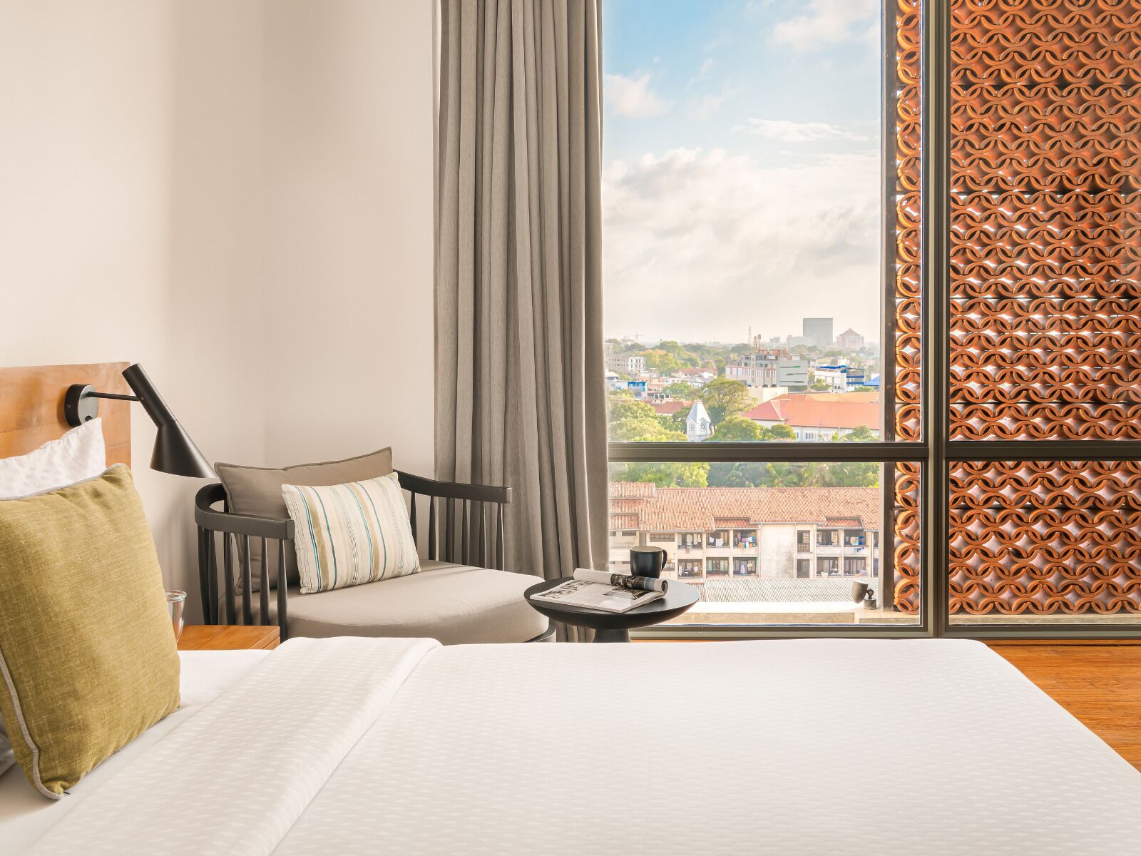 Habitación de hotel con un ventanal grande, una cama, una silla, una mesita con una taza y vistas a un paisaje urbano.