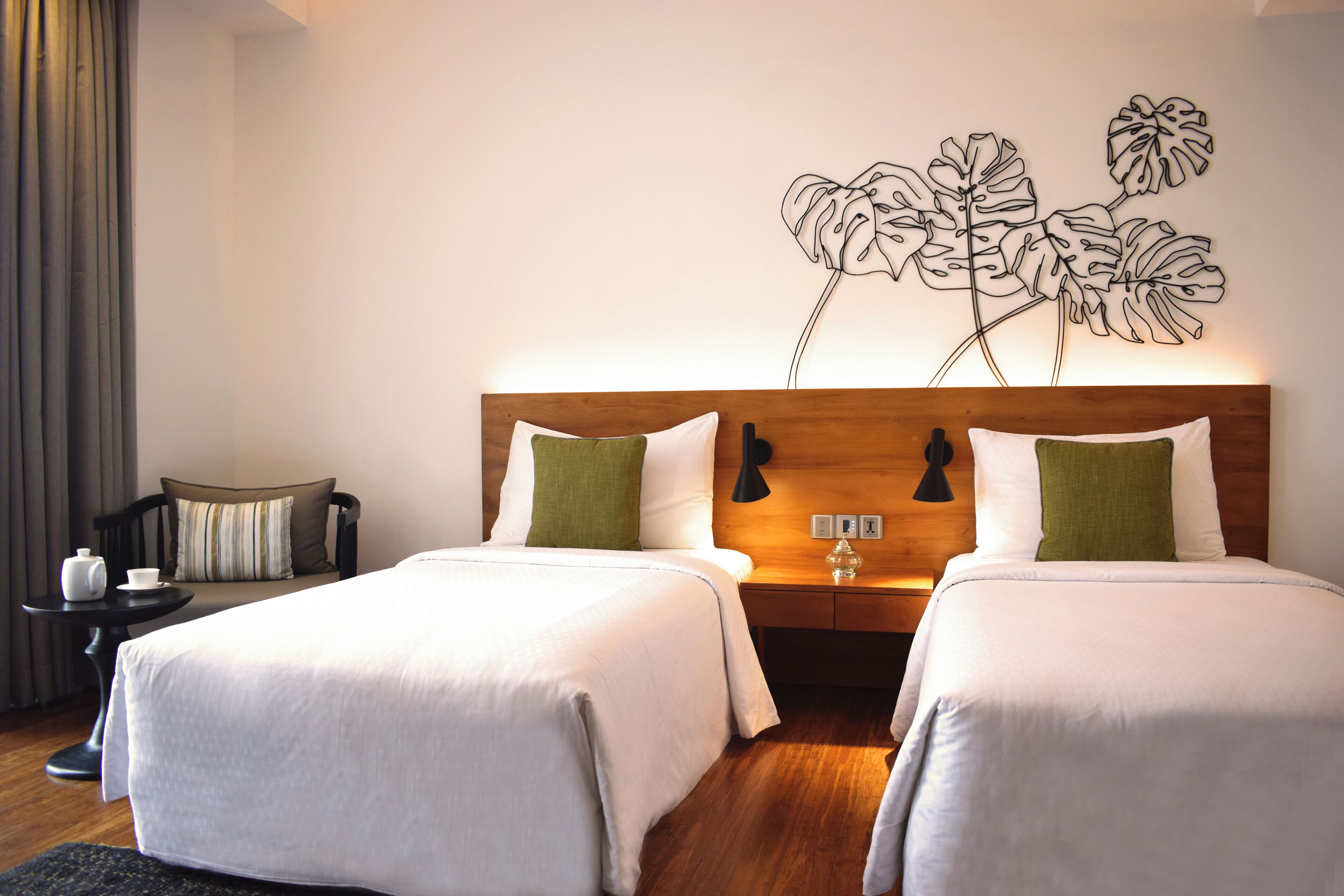 A hotel room with two beds, a wooden headboard, a wall art of abstract leaves, a small table with a cup and saucer, and a chair.