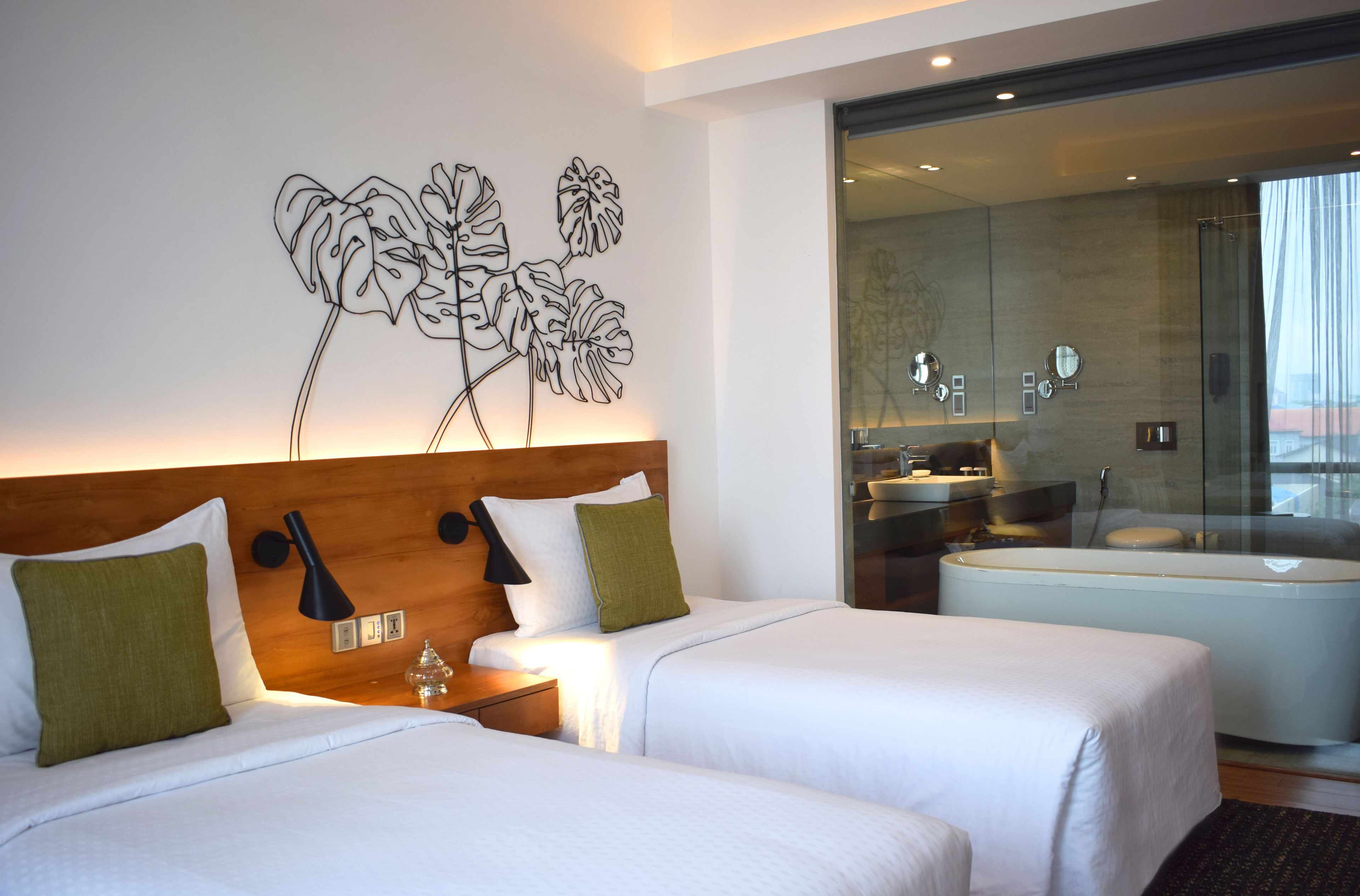 A hotel room with two beds, a wooden headboard, a wall art of abstract leaves, a bathroom with a bathtub, and a glass shower enclosure.