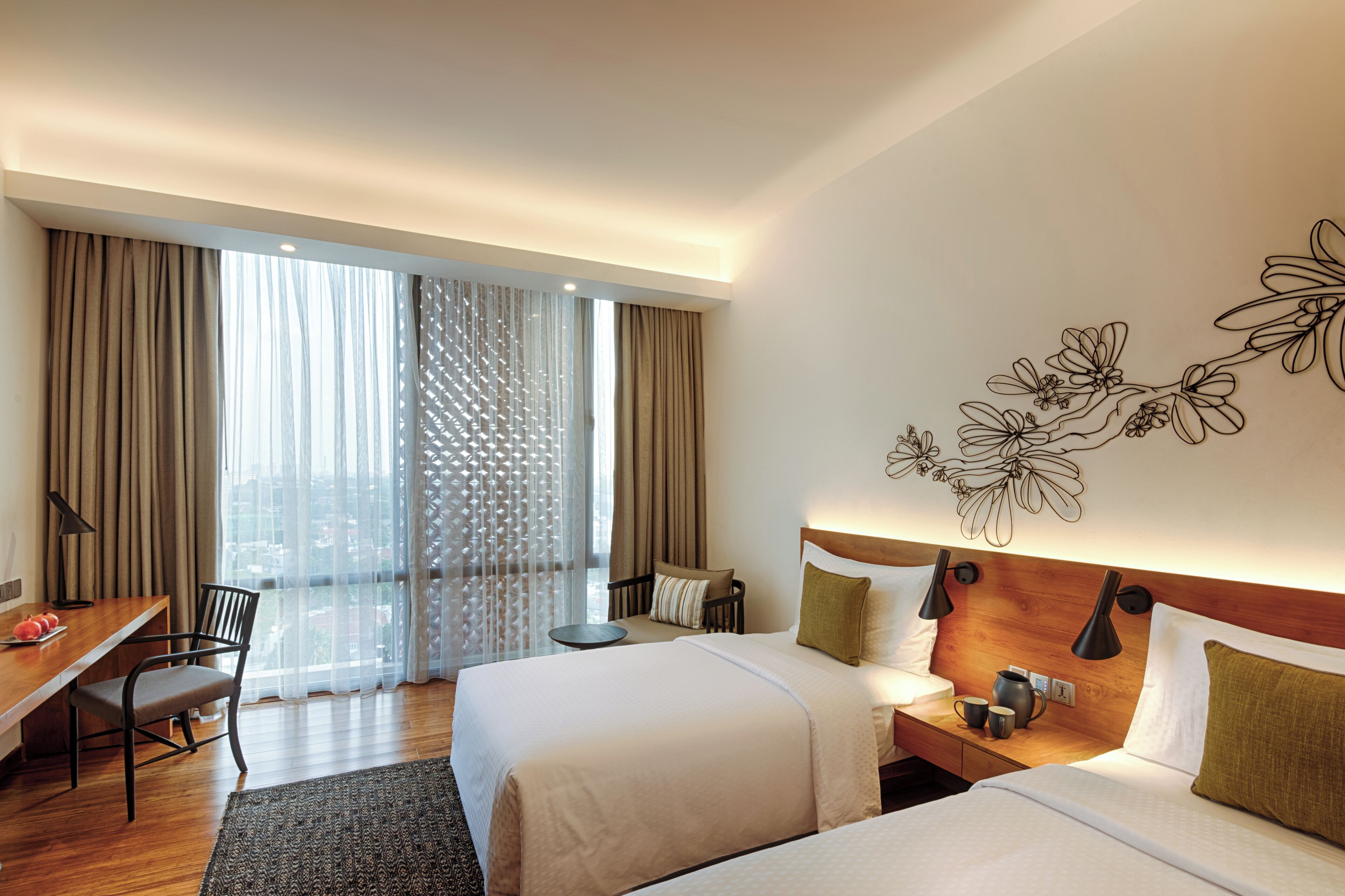 A hotel room with two beds, a desk, a chair, a small table, and a view of the city through large windows.