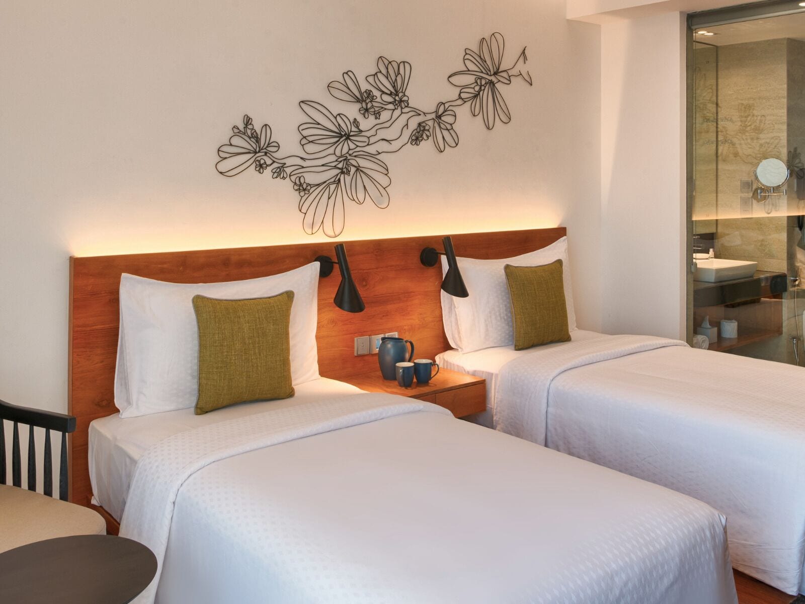 A hotel room with two beds, a wooden headboard, a wall-mounted lamp, a nightstand with a coffee pot, and a bathroom visible through a glass partition.
