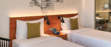 A hotel room with two beds, a wooden headboard, a wall-mounted lamp, a nightstand with a coffee pot, and a bathroom visible through a glass partition.