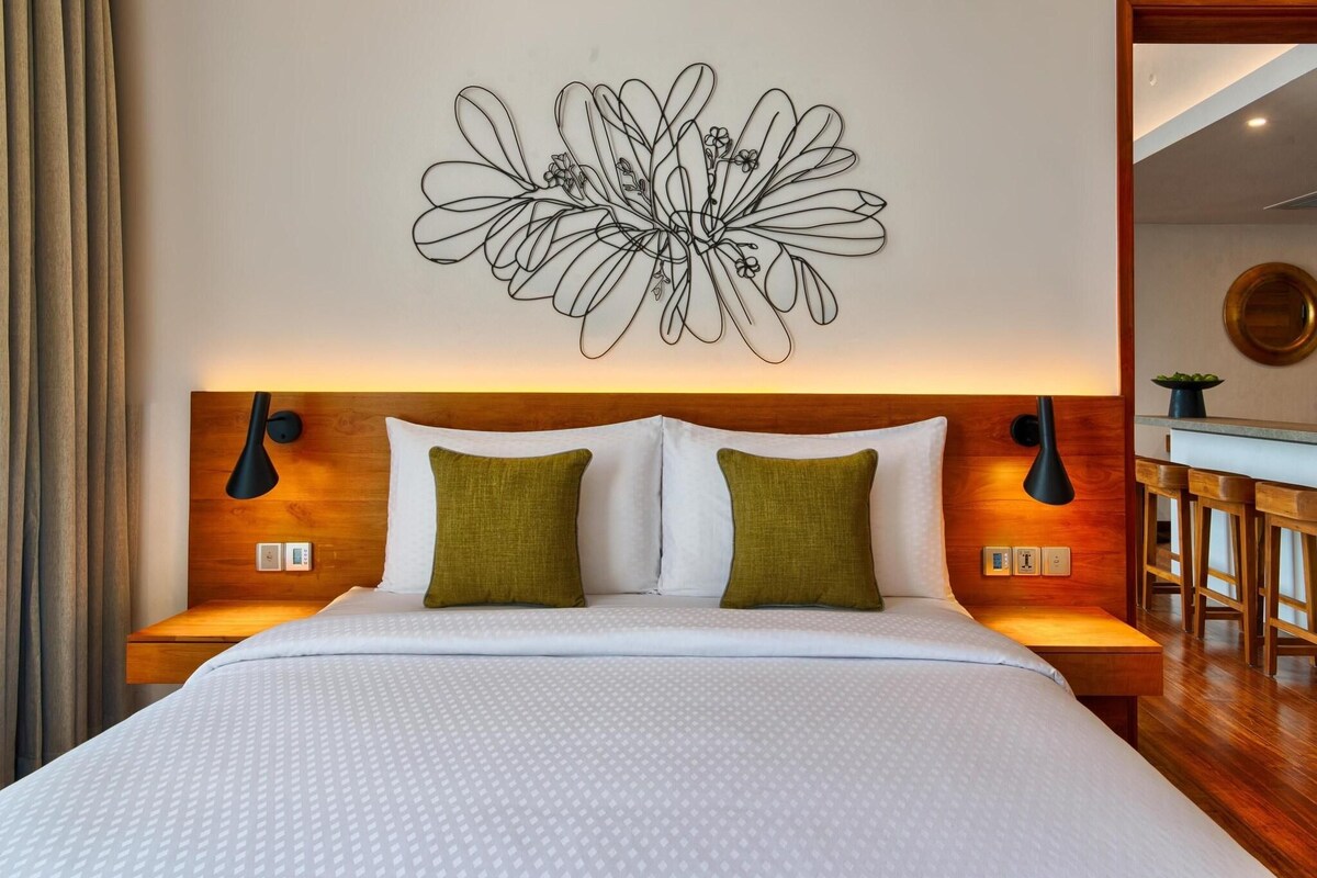 Una cama bien hecha con disposición de almohadas blancas y verdes, un cabecero de madera y una obra de arte abstracta colgada en la pared.
