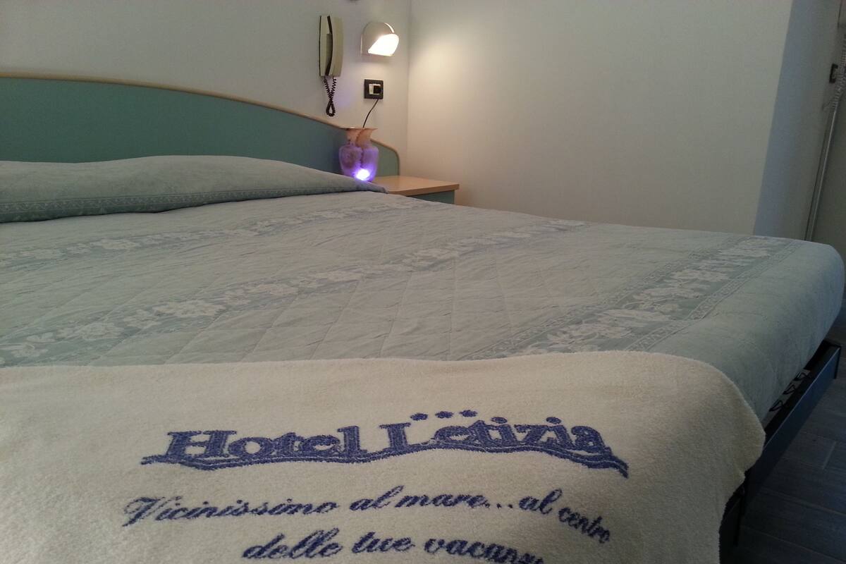 Ein Bett mit weißer Bettdecke und blauer Stickerei. Ein Nachttisch mit Lampe und Telefon.