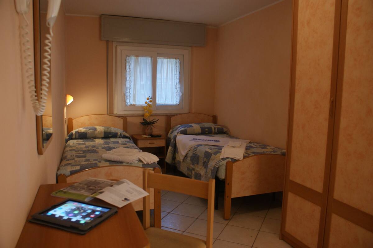 Ein kleines Schlafzimmer mit zwei Betten, einem Nachttisch mit Lampe, einem Sessel und einem Schreibtisch mit Tablet.