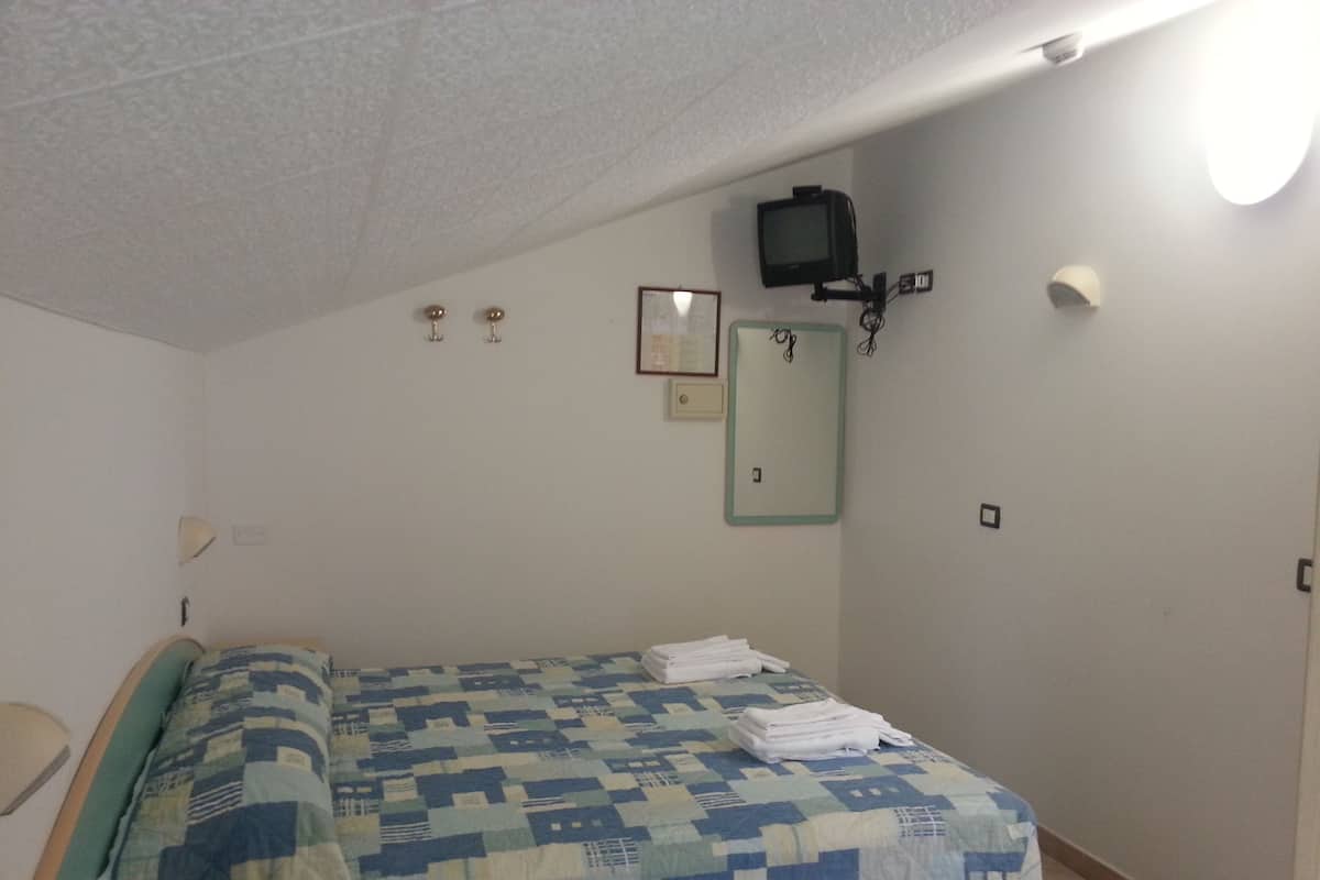 Ein kleines, schlichtes Zimmer mit einem Bett, einem an der Wand befestigten Fernseher und einem kleinen Tisch mit Stuhl.