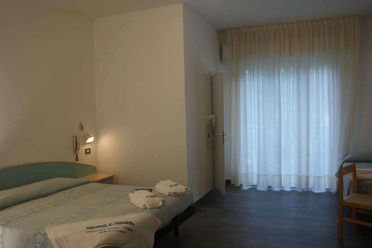 Ein Hotelzimmer mit einem Bett, einem Nachttisch mit Lampe, einem Sessel und einem Fenster mit transparenten Vorhängen.