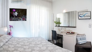 A hotel room with a bed, a desk, a chair, a small table with a vase and a bottle, and a bag on a stool.
