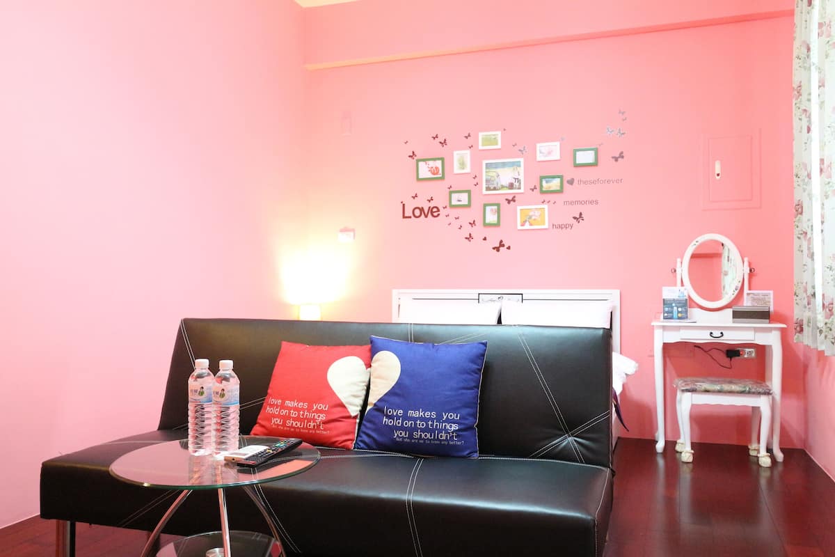A pink-themed room with a black leather sofa, two heart-shaped pillows, a glass table with water bottles, a small white desk with a mirror, and a wall decorated with framed photos and butterflies.