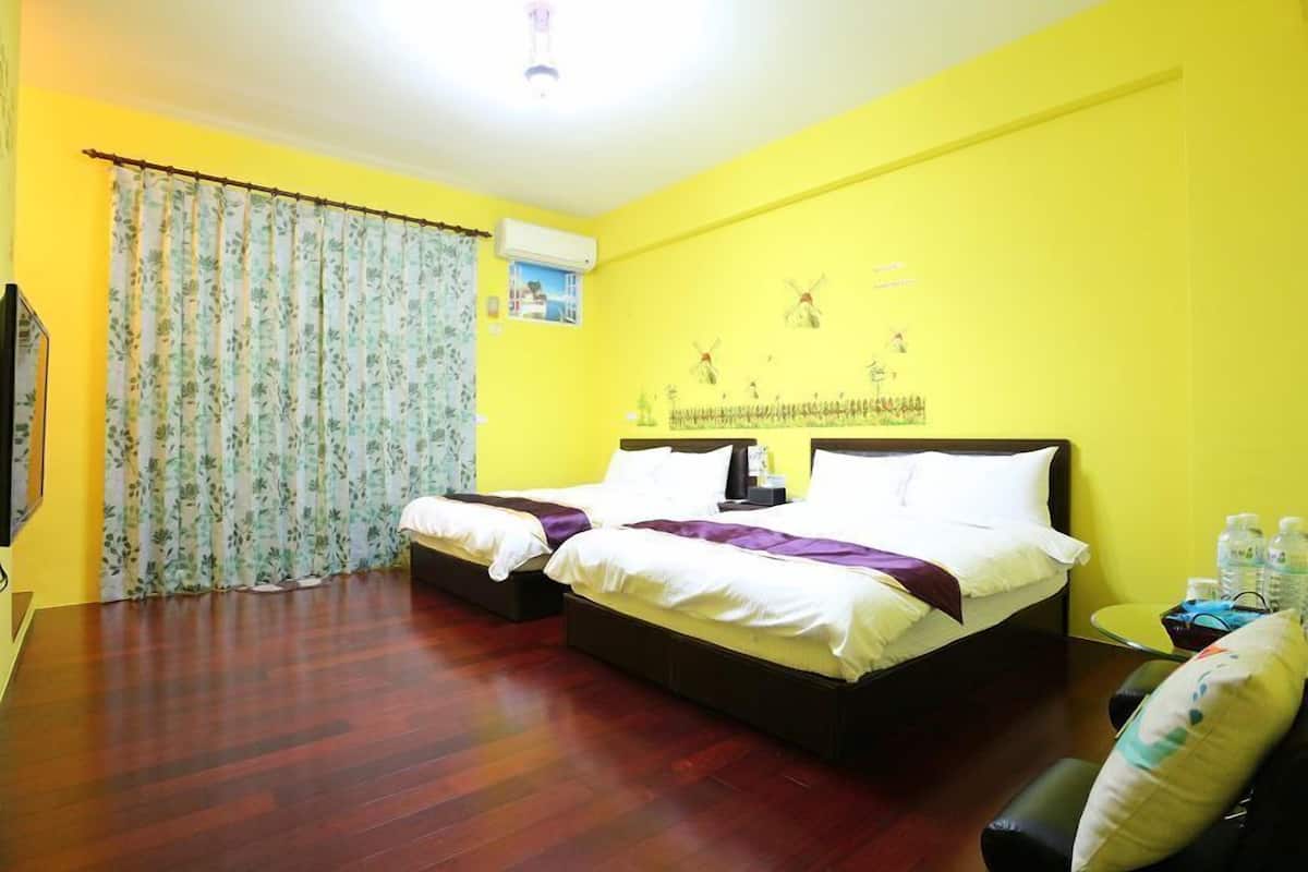 A hotel room with two beds, a wooden floor, a yellow wall, and a window with floral curtains.