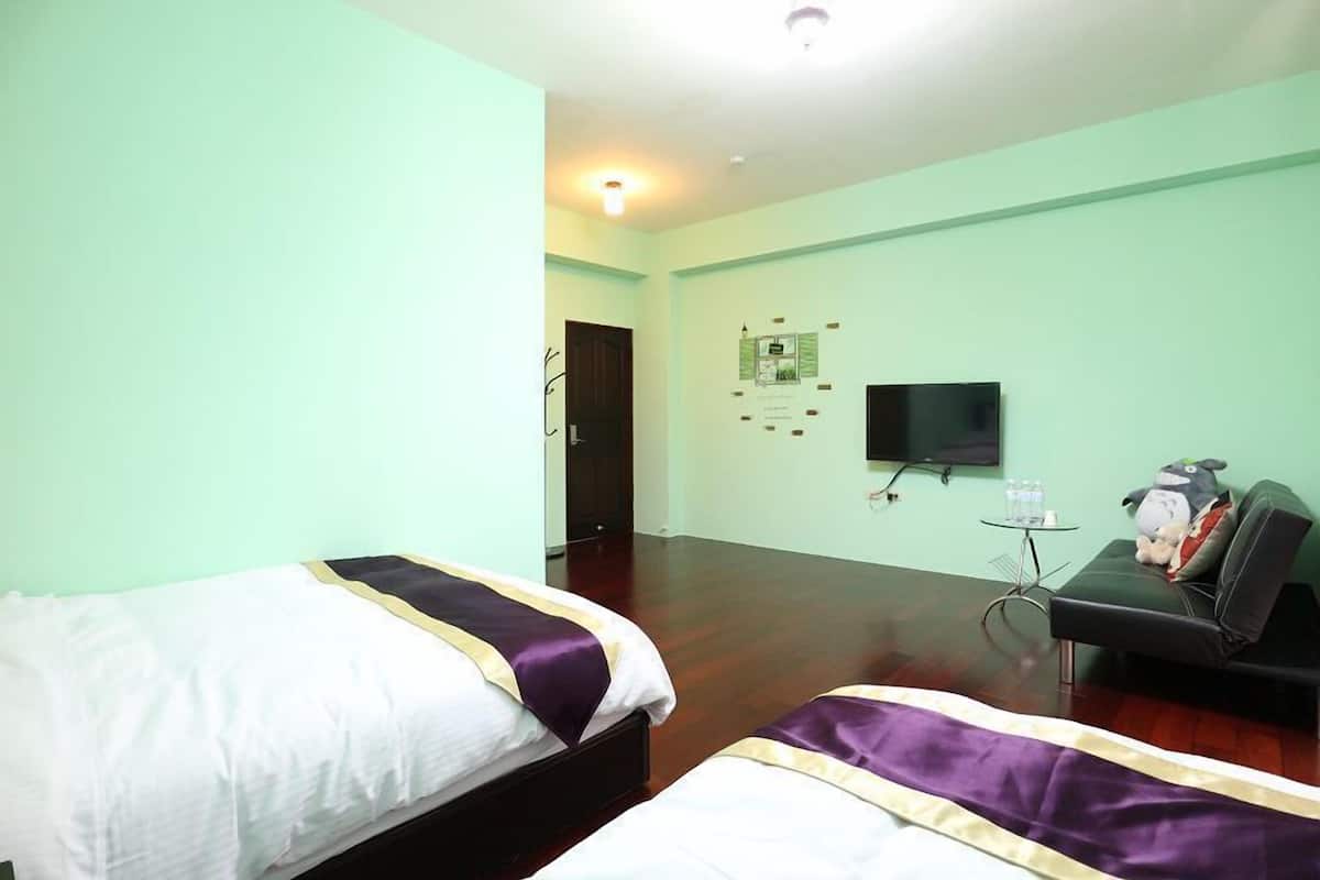 A hotel room with two beds, a flat-screen TV, a small table with a water bottle, and a couch with a stuffed toy.