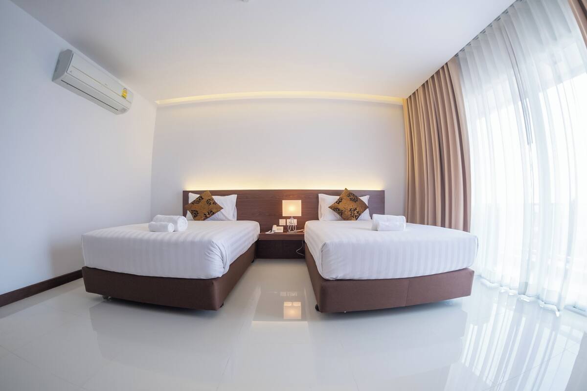 A hotel room with two beds, a nightstand, a lamp, and a large window with curtains.