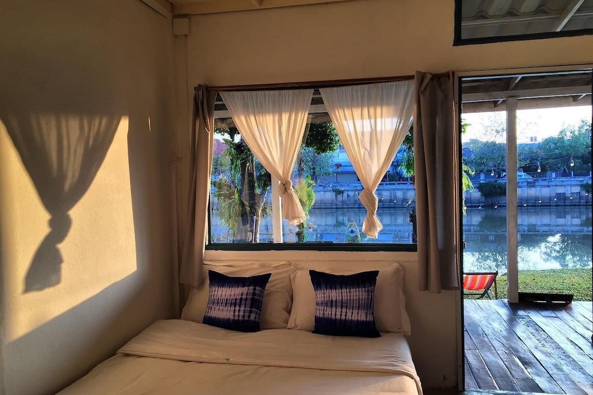 A bedroom with a bed, two pillows, a window with sheer curtains, and a view of a water body and greenery outside.
